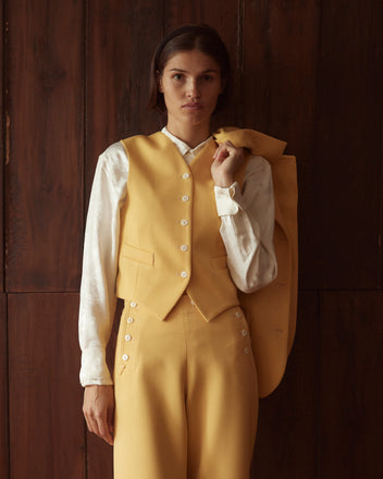 A person wearing the Riding Twill Waistcoat in yellow, with a vintage flair, holds a matching jacket while standing against a wooden backdrop.