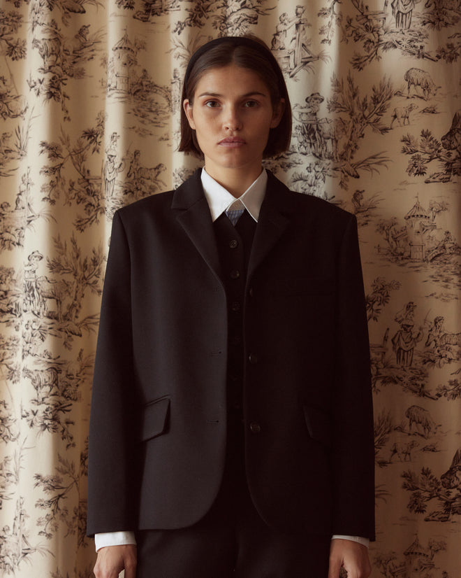 Dressed in a sophisticated Riding Twill Lillian Jacket - Black, a person stands gracefully against a patterned curtain backdrop.