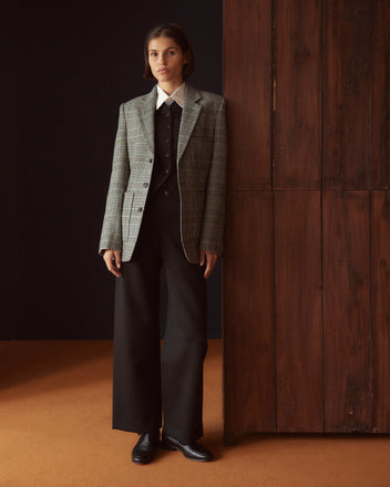 A person wearing the Blue Mountain Suit Jacket - 36 paired with wide-leg trousers made from 1960s suiting fabric stands by a wooden wall in a dimly lit room.