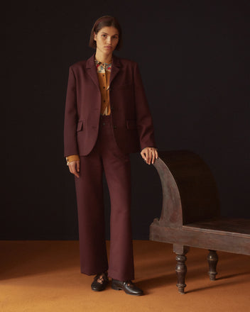 A person in a maroon suit and yellow shirt, paired with Riding Twill Sailor Trousers - Burgundy, stands beside a vintage bench on an orange floor.