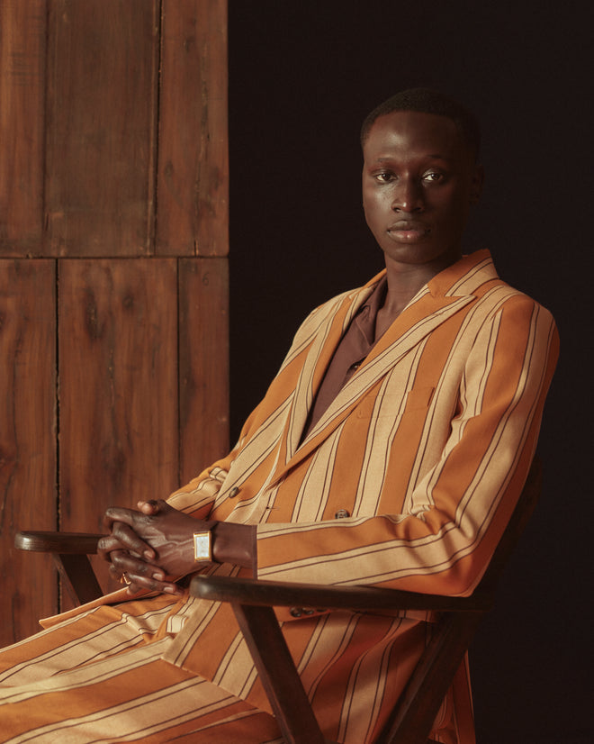 A person wearing the Awning Stripe Double-Breasted Suit Jacket, crafted from midcentury yardage, leans against a wall in a softly lit room.