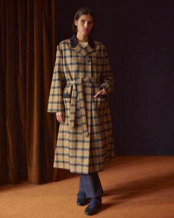 A person stands on an orange carpet near brown drapes, their attire a testament to timeless style—a long Equestrian Locke Coat crafted from British mill windowpane twill, elegantly belted at the waist.