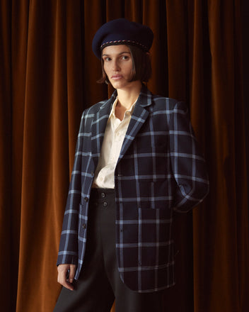 A person stands in front of brown curtains, wearing the unique Criterion Plaid Suit Jacket - 36, crafted from 1960s suiting fabric, paired with a crisp white shirt and a stylish navy beret.