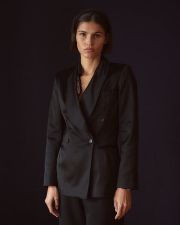 A person wearing the Sheen Suit Jacket, designed with a satin weave and highlighted by a sleek shawl collar, stands against a dark background, looking directly at the camera.