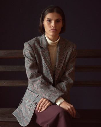 A person with short dark hair wearing the Speckled Feather Suit Jacket - 36 sits on a wooden bench against a dark background.