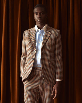 A person with long hair is wearing the Marston Check Suit Jacket paired with a white shirt made from 100% merino and brown loafers, standing against a light yellow wall.