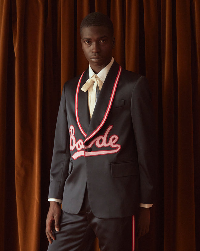 A person wearing the Major League Suit Jacket, a stylish dark suit with red trim and embroidered text, featuring a classic shawl collar, stands against a brown curtain backdrop.