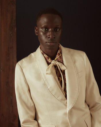 A person in the Roping Shawl Collar Tuxedo Jacket and brown shoes stands against an off-white wall, exuding elegance with its shimmering cream-colored twill fabric accents.