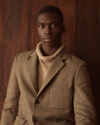 Against a wooden background, a person is wearing the Macnamara Suit Jacket - 40 in brown over a turtleneck, evoking classic 1960s suiting fabric with its 100% wool composition.