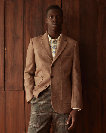 A person in a Carbon Print Suit Jacket - 40 and plaid pants stands confidently against a wooden wall, showcasing timeless style with suiting fabric that exudes elegance.