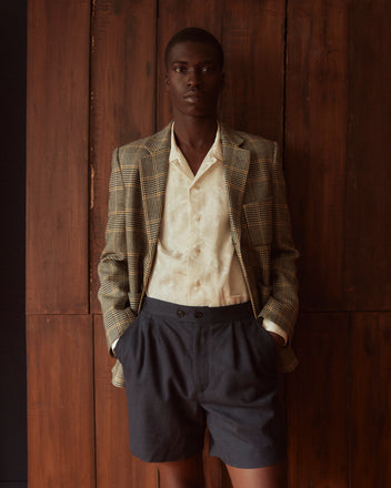 A person in a plaid blazer, white shirt, and Herringbone Wool Shorts - 32 with side-buckle waist adjusters stands against a wooden background.