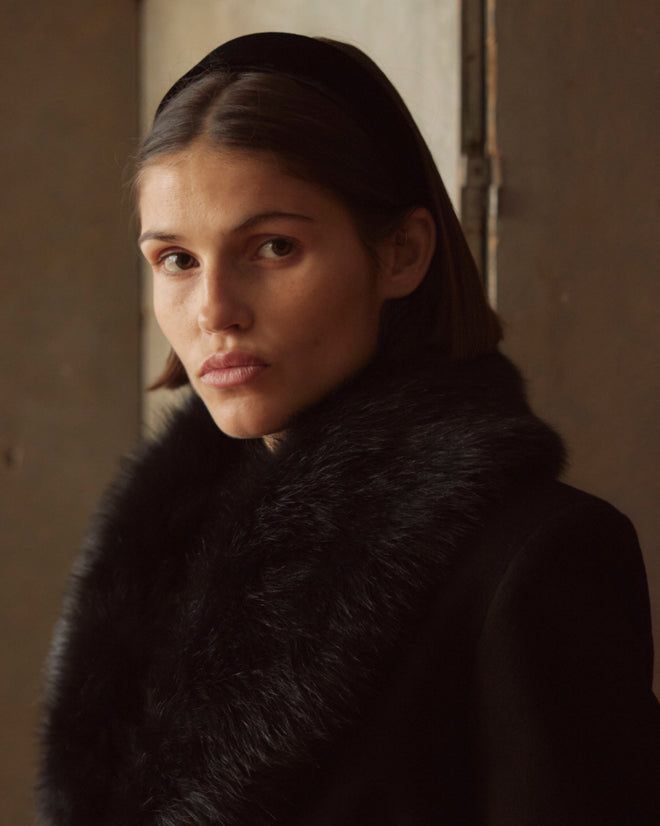 A woman with short hair dons the Arena Coat, featuring a black wool design with a fur collar, looking directly at the camera.