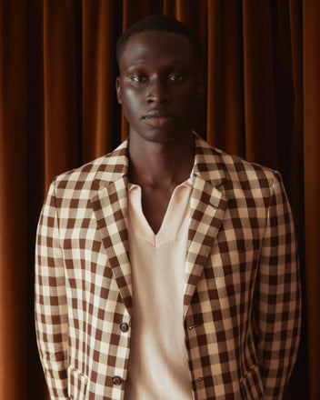 A person stands against a light-colored wall, adorned in a Gilbert Check Single-Breasted Suit Jacket made from deadstock fabric, featuring a brown and white checkered pattern reminiscent of the 1950s. Their outfit is completed with matching trousers and brown shoes.