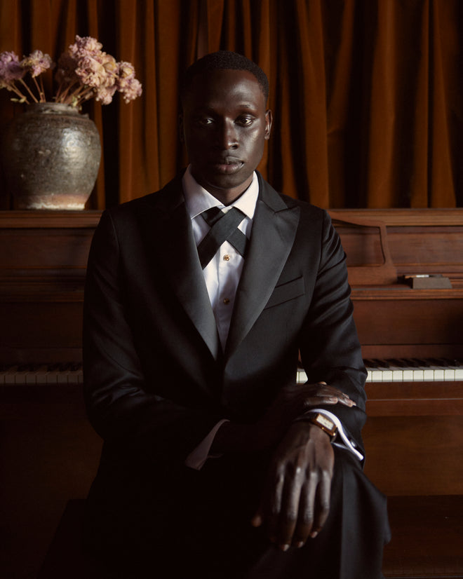 A person in a Barathea Peak Lapel Tuxedo Jacket sits in front of a piano, with a vase of flowers gracing the background.