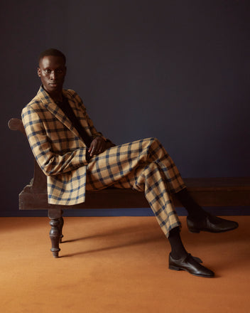 A man in a plaid suit sits on a bench, his Equestrian Trousers with side-buckle waist adjusters providing a striking contrast against the dark blue wall and vivid orange floor.