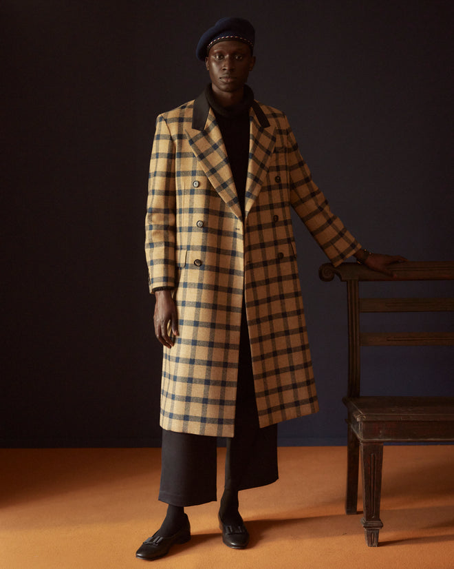 A person wears the Equestrian Mitsy Coat, a double-breasted plaid wool coat with black lapels, crafted from classic British mill fabric, standing stylishly against a tiled background.