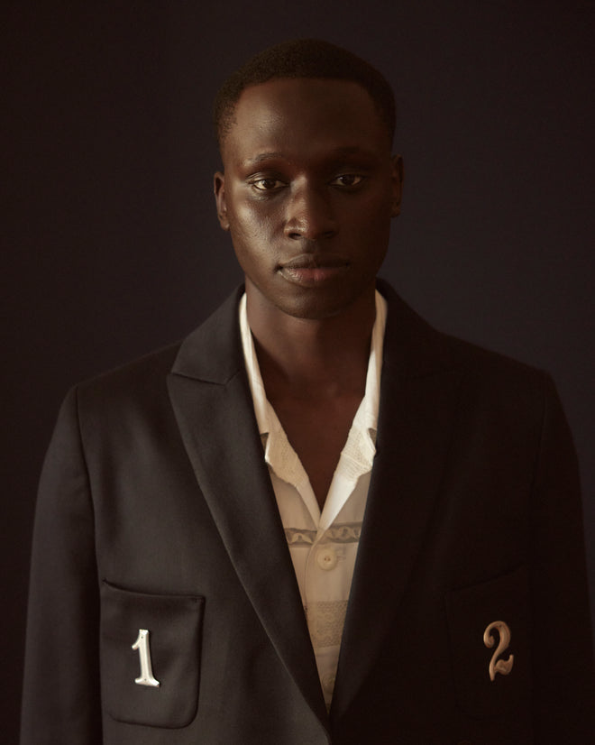 Dressed in the Numerals Suit Jacket, a person embraces the classic 1940s style with barathea wool and numbered pockets, all set against a dark backdrop.