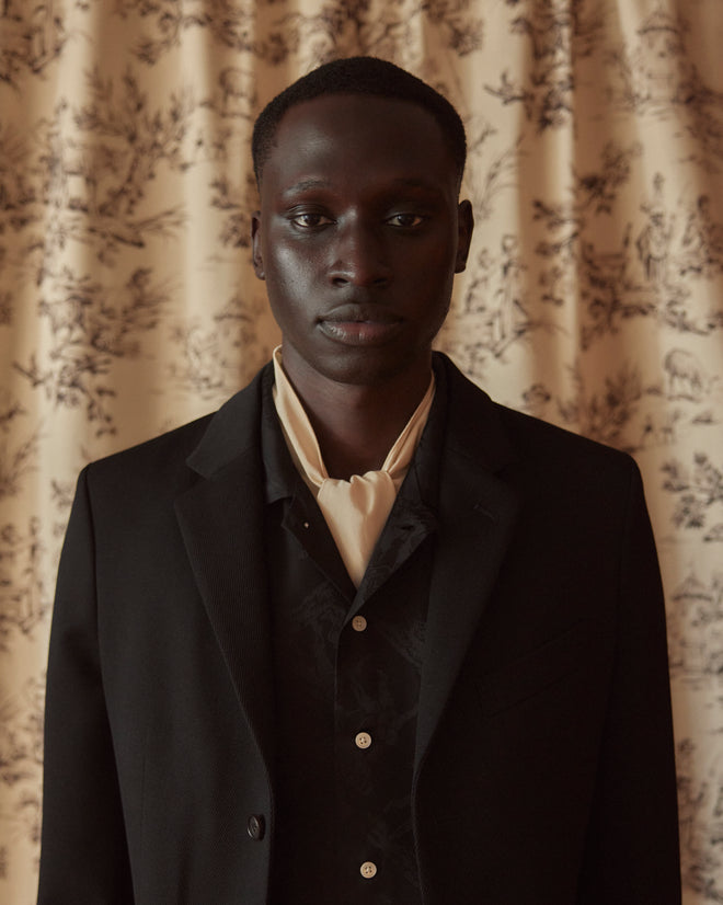 A person wearing the Riding Twill Suit Jacket in black stands in front of a patterned curtain, looking towards the camera.