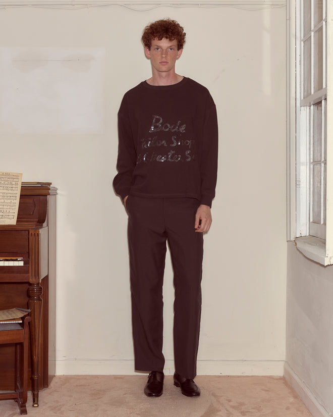 A man wearing the Tailor Shop Crewneck - Black stands by a piano near a window in a softly lit room, bringing vintage charm to the scene.