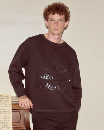 A young man with curly hair wears the Tailor Shop Crewneck - Black, standing by a piano with sheet music on top, capturing the vibe of a vintage advertisement photo.