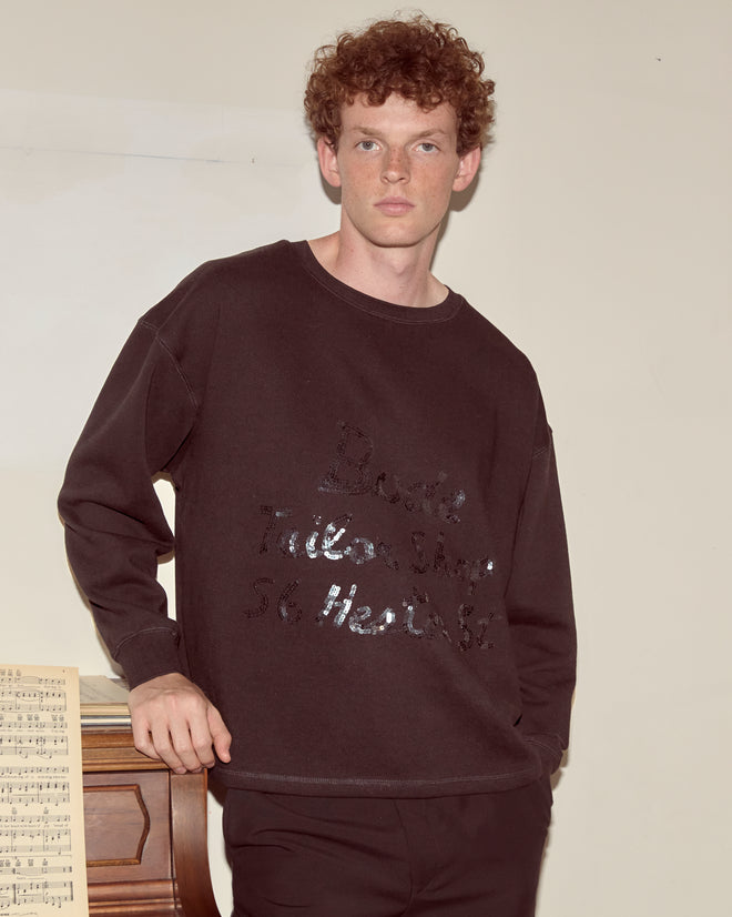 A young man with curly hair wears the Tailor Shop Crewneck - Black, standing by a piano with sheet music on top, capturing the vibe of a vintage advertisement photo.