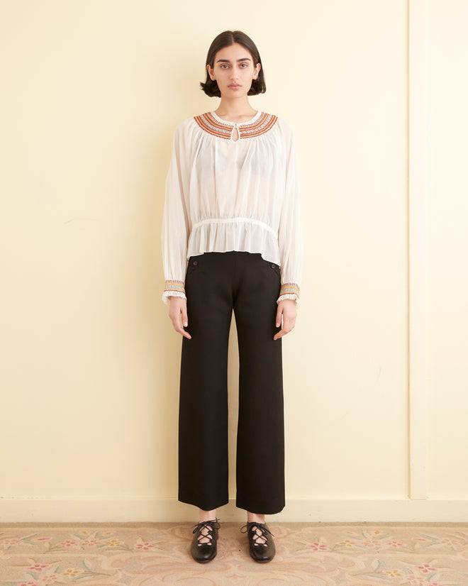 A person stands against a plain, light-colored wall, dressed in the Embroidered Georgette Top with red smocking embroidery, paired with black wide-legged pants and black shoes.