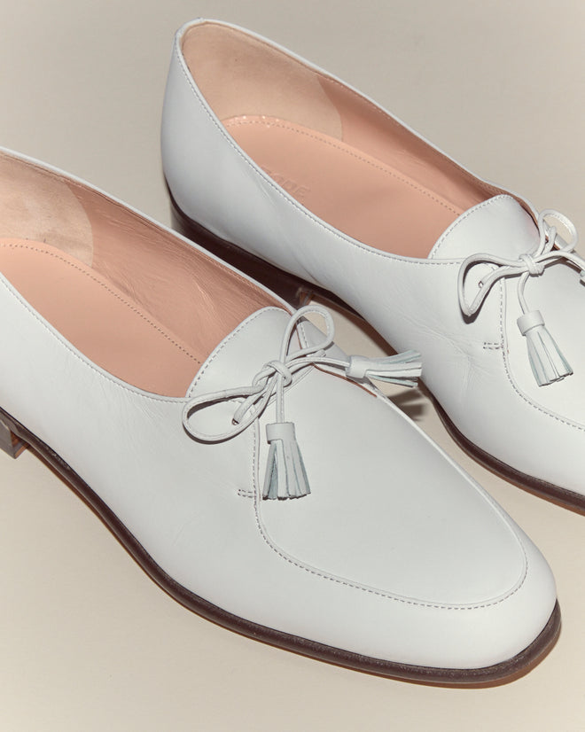 The Tassel Shoes - White feature pink interiors, tassel details, and low brown heels for vintage loafer style. Made from premium calf leather, they stand out stylishly against a light background.
