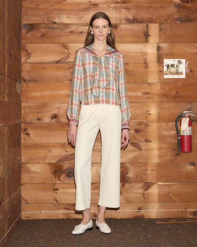 A woman wearing the Canyon Plaid Blouse, which hints at 1920s style, and white pants stands indoors beside a wooden wall, a fire extinguisher, and a cow picture.