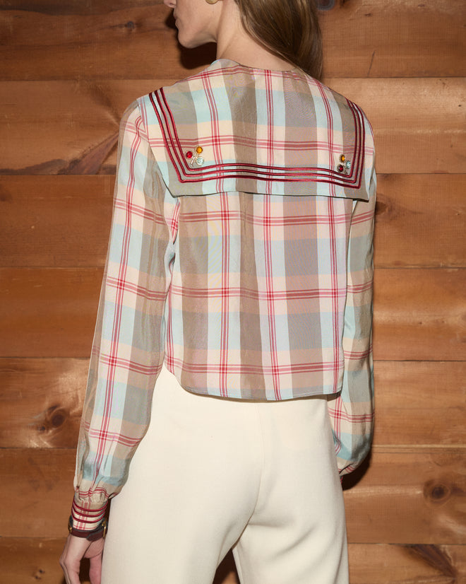 A woman stands against a wooden wall, facing away from the camera, wearing the Canyon Plaid Blouse with a sailor collar and cream pants in a 1920s-inspired style.