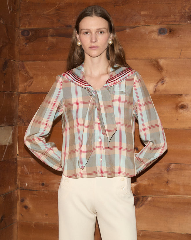 Wearing the Canyon Plaid Blouse with its distinctive 1920s-inspired sailor collar and paired with cream pants, a woman stands against a wooden wall, gazing at the camera.