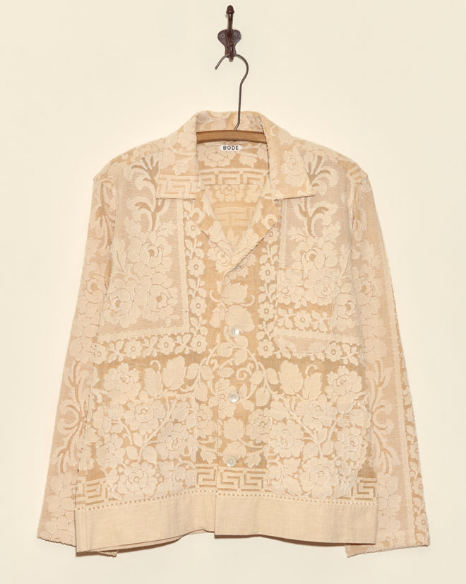 The Sandstone Key Overshirt in size S, featuring a unique beige jacquard lace with a delicate floral pattern, hangs on a hook against a light-colored wall.