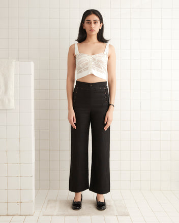 In a tiled room, a person is wearing a stylish Tennis Lace Tank and black high-waisted pants, elegantly paired with black shoes.