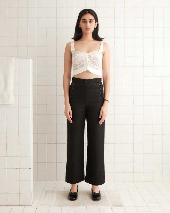 In a tiled room, a person is wearing a stylish Tennis Lace Tank and black high-waisted pants, elegantly paired with black shoes.