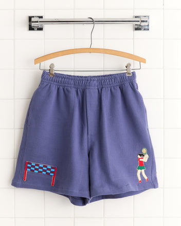 Tennis Player Shorts in purple feature tennis-themed embroidery of a player and net. Inspired by classic Canadian rugby shorts, they bring playful style to any sporty wardrobe and hang on a wooden hanger.