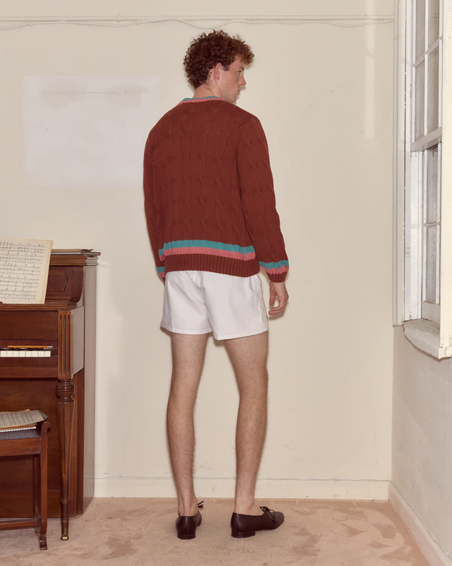 A person in a brown Tennis Sweater stands facing a wall near a piano in a softly lit room.