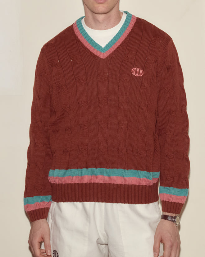 Man wears the Tennis Sweater - Brown, a maroon cable-knit with green and pink trim, paired with white pants—classic knitwear inspired by Bode’s signature style.