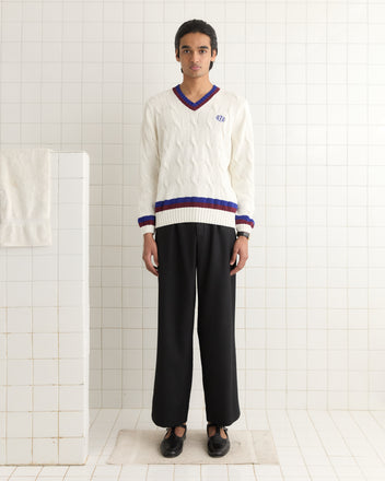 A person wearing a Tennis Sweater with navy and burgundy stripes stands on a rug in a tiled room, paired seamlessly with black pants.