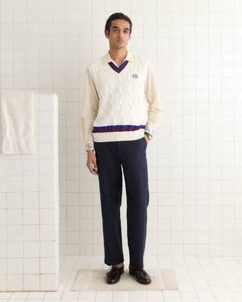 A person wearing a Tennis Vest and navy trousers stands in a white tiled room with a towel on a rail.