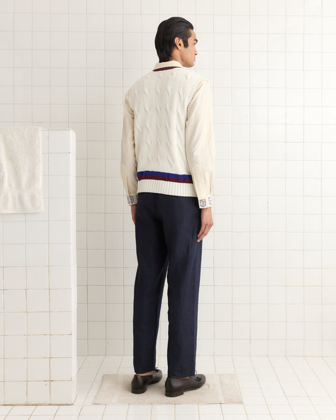 A man in a cream Tennis Vest and dark pants stands in a tiled room, back to the camera, radiating understated elegance.