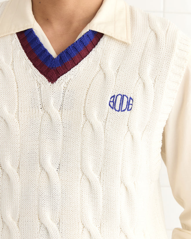 A person is wearing the Tennis Vest, a cream cable-knit with blue and burgundy trim and Bode logo embroidered on the chest.