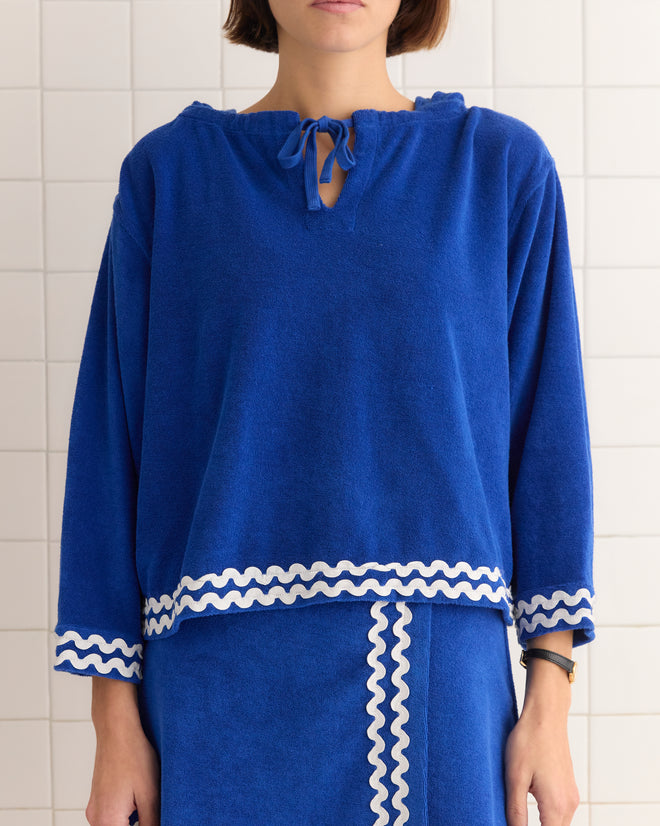 A person wearing the Terry Trim Hoodie, a blue terrycloth outfit with white wavy trim, stands against a tiled wall.