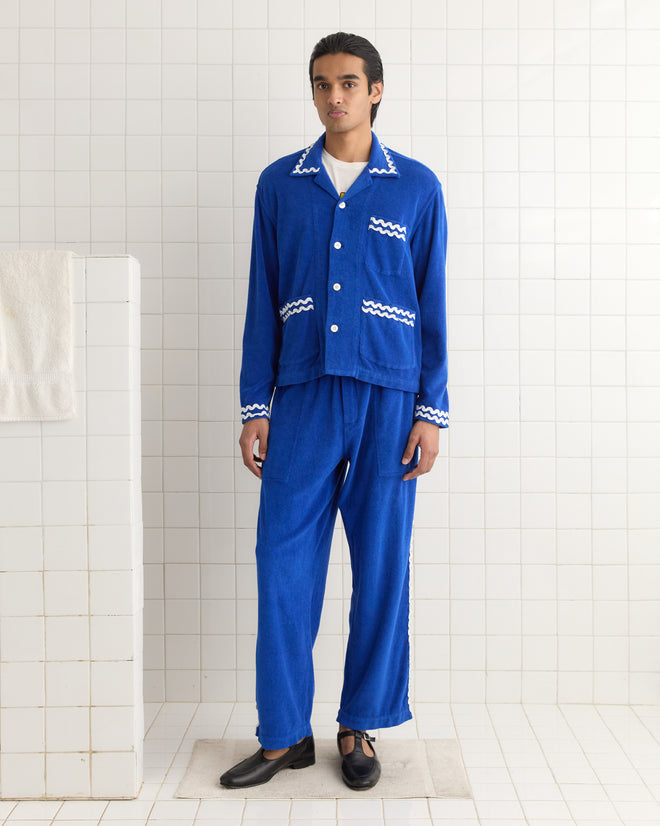 A person in blue pajamas with a Terry Trim Overshirt featuring white zigzag pattern stands in a tiled room, next to a hanging terrycloth towel on the left.