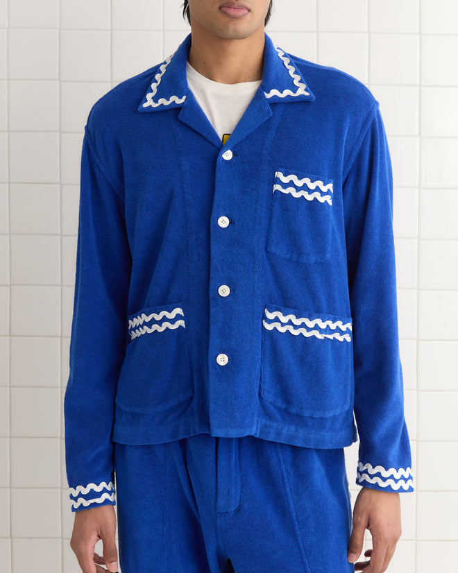 A person in a Terry Trim Overshirt, featuring blue with white wavy trims reminiscent of a 1980s-inspired French tracksuit, stands against a tiled wall.