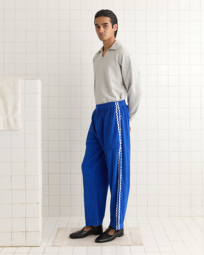 A person wearing a gray top and Terry Trim Track Pants stands on a mat in a tiled room, hands behind their back, facing slightly sideways.