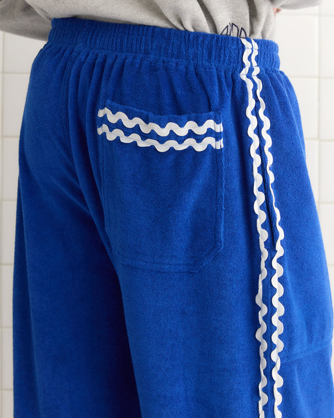 Someone wears Terry Trim Track Pants, featuring a blue terrycloth design with wavy white trim on the side and pocket, standing against a tiled backdrop in a style that echoes vintage 1980s tracksuits.