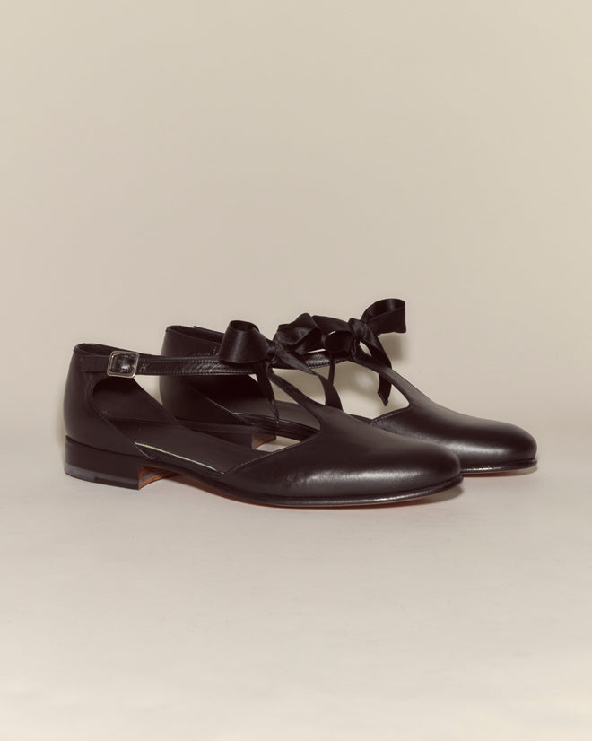 The Theater Shoes - Black are black leather flats featuring cut-out sides, ankle straps, and bows, evoking classic T-strap style on a neutral background.