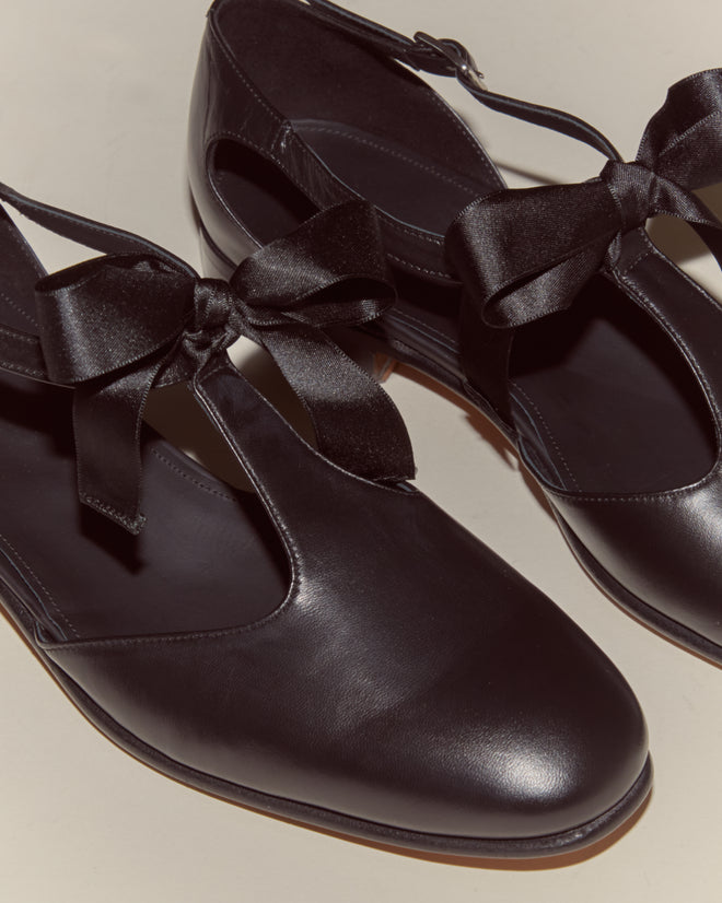 The Theater Shoes - Black are black leather T-strap flats with satin ribbon bows and ankle straps, displayed on a light surface.