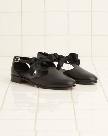 The Theater Shoes - Black, crafted from leather and adorned with wide black bows and side buckles, are displayed on white tiled flooring.