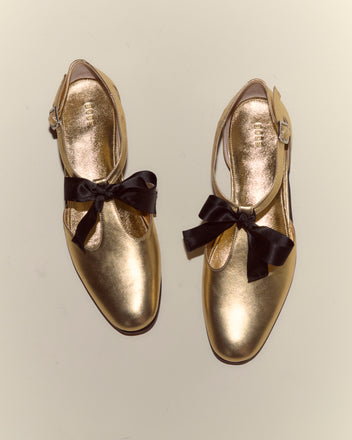 Theater Shoes - Gold feature a shiny metallic finish, black bows, and buckled T-straps, inspired by midcentury dancing shoes; shown from above on a plain background.