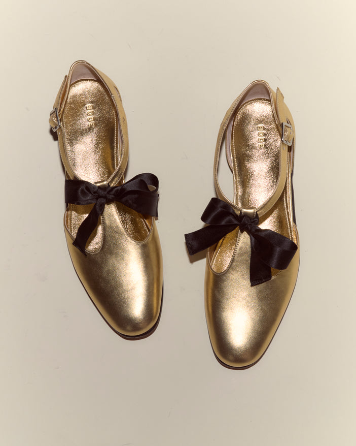 Theater Shoes - Gold feature a shiny metallic finish, black bows, and buckled T-straps, inspired by midcentury dancing shoes; shown from above on a plain background.